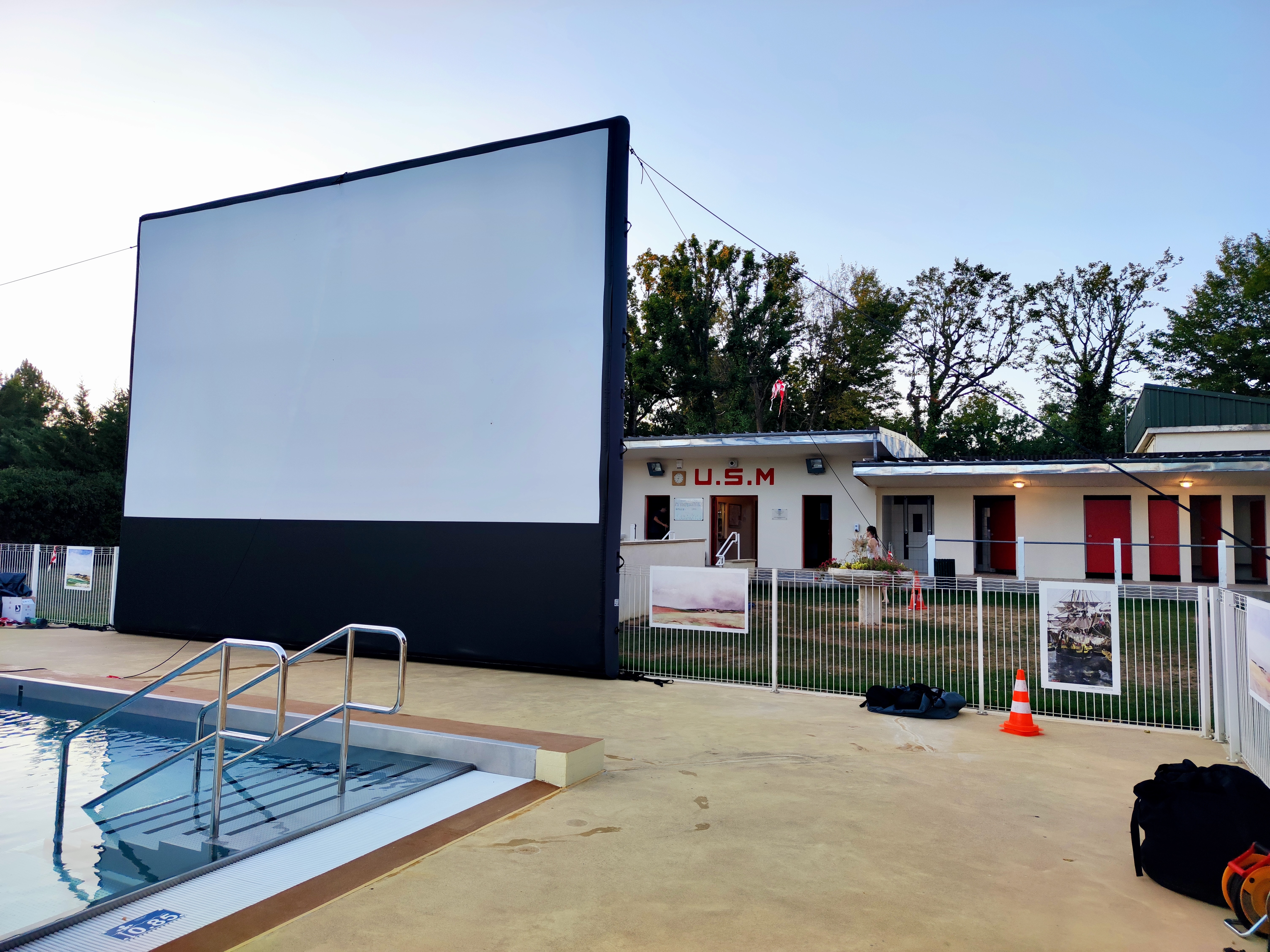 Grand écran gonflable noir et blanc installé au bord d'une piscine municipale, devant un bâtiment portant l'inscription U.S.M.
