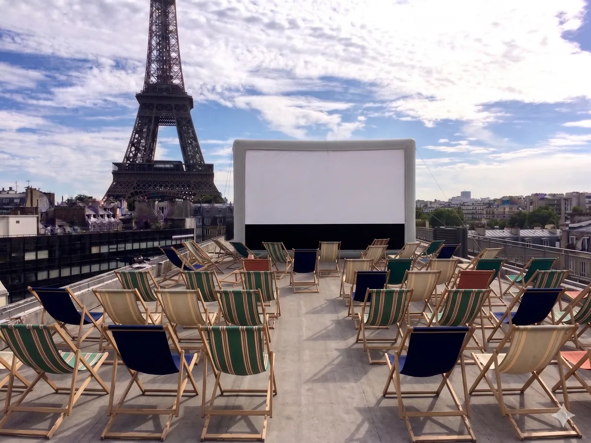 Écran gonflable blanc installé sur une terrasse en hauteur à Paris, entouré de chaises longues colorées vides face à la Tour Eiffel visible en arrière-plan sous un ciel dégagé.