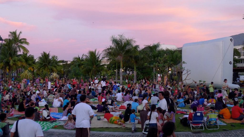 Une foule nombreuse assise sur des nattes et couvertures attend devant un écran gonflable blanc, sous un ciel rose de crépuscule bordé de palmiers.