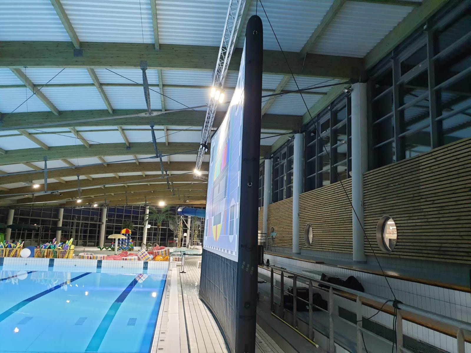 Vue de profil d'un écran gonflable noir suspendu sous la charpente d'une piscine couverte, avec mire de calibration affichée et bassin bleu en contrebas.