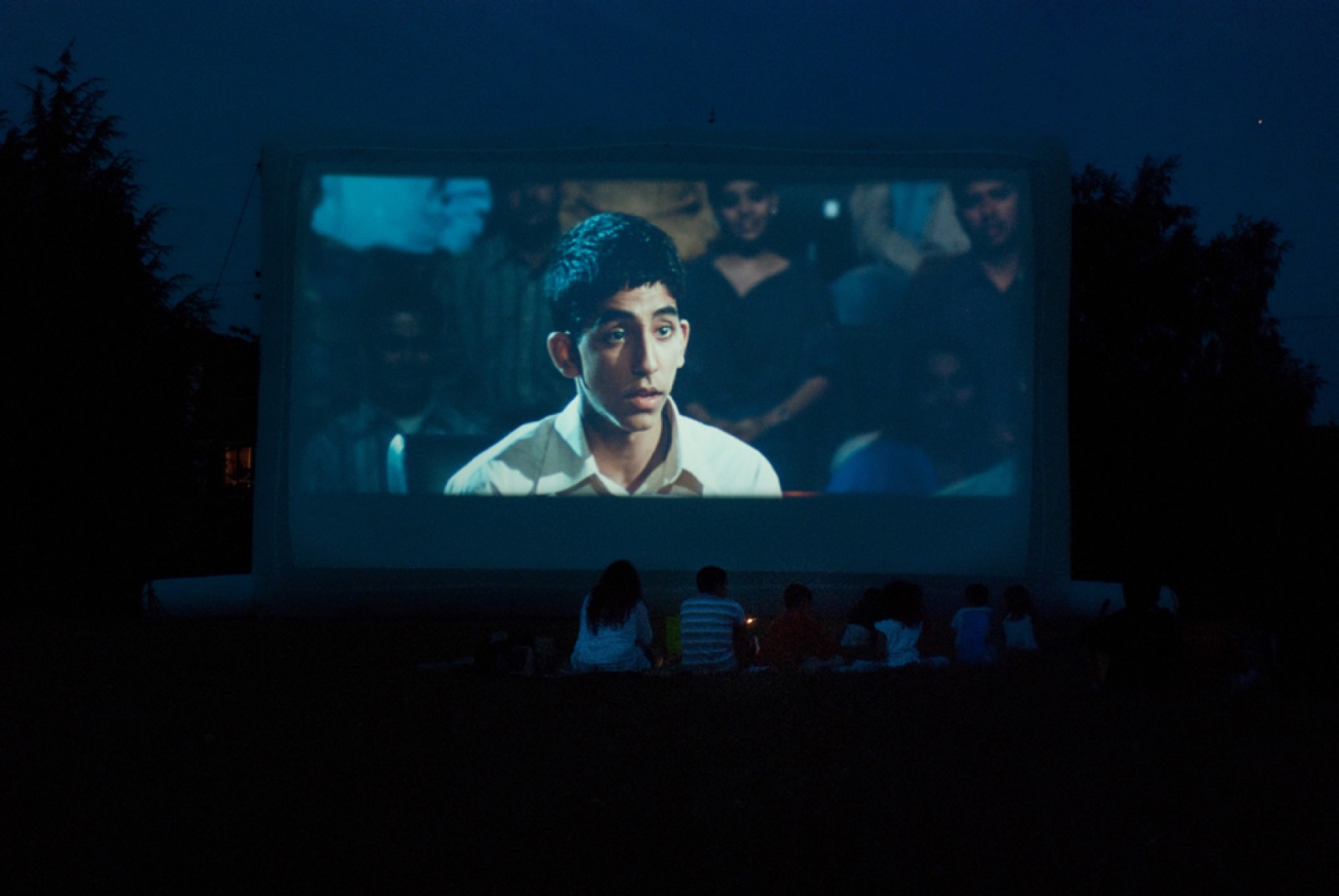 Open-air movie screening on an inflatable screen; the audience settles in front of the screen as night falls
