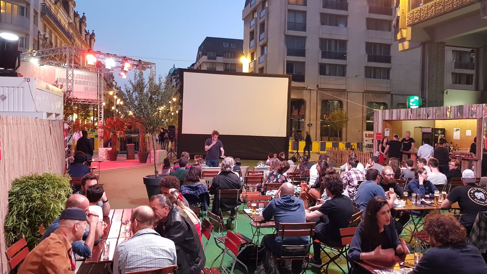 Un écran gonflable installé sur une place urbaine accueille un public nombreux attablé en terrasse à la tombée de la nuit.