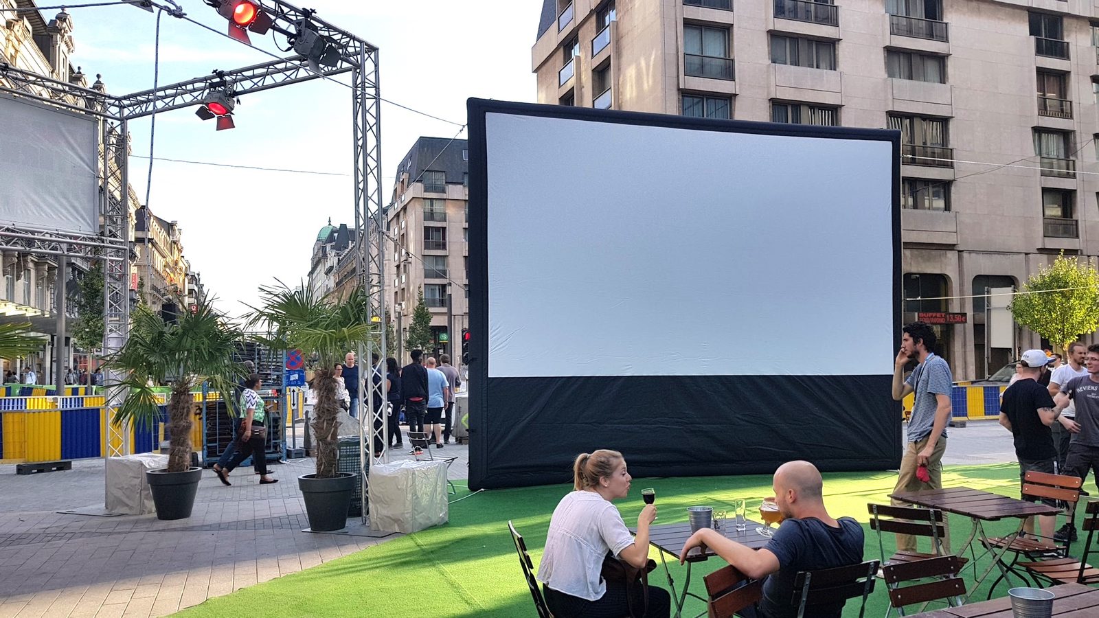 Un écran gonflable installé sur une place urbaine avec des personnes attablées en terrain buvant un verre au premier plan.