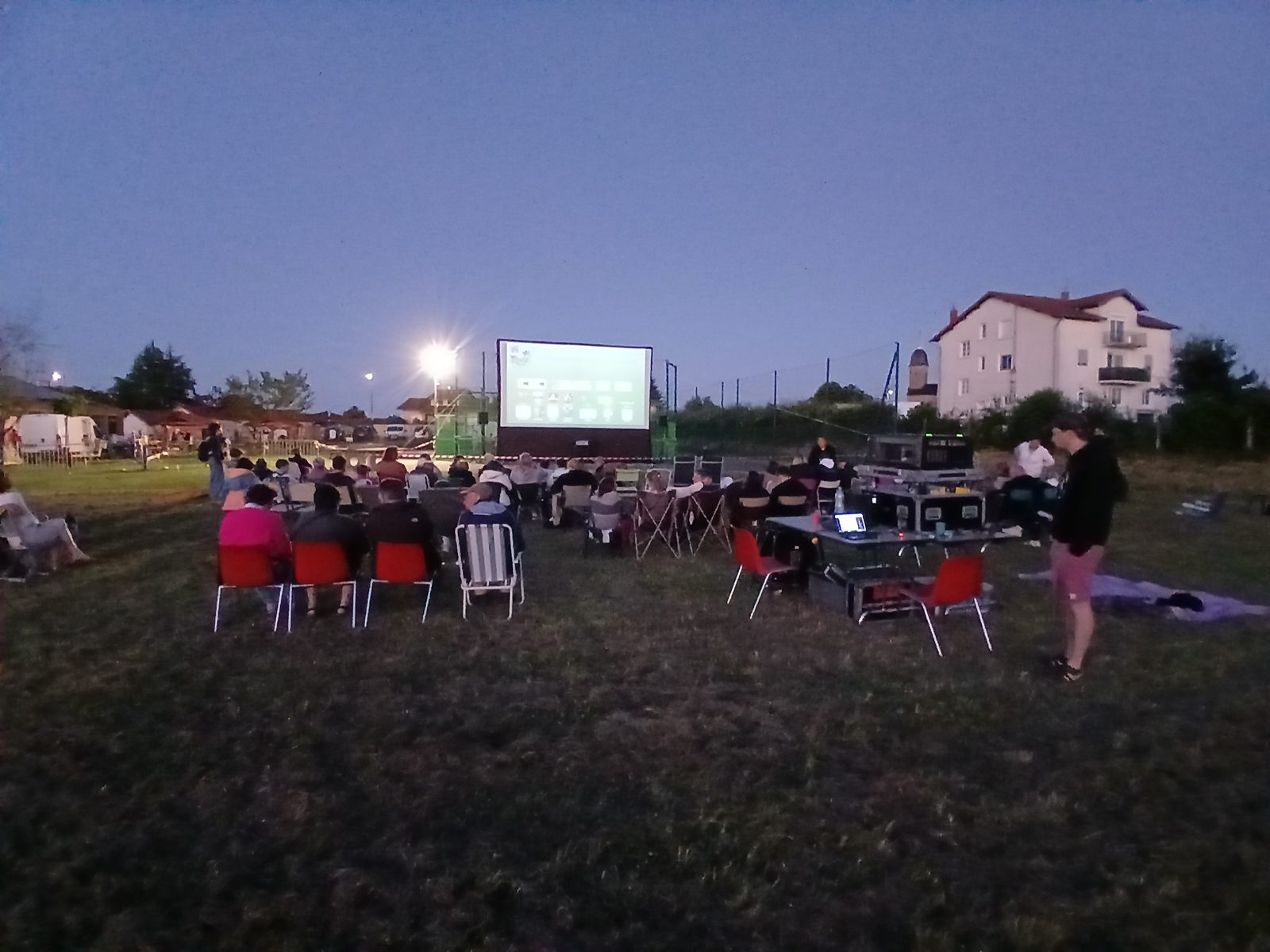 Un écran gonflable étanche diffuse un film au crépuscule devant un public assis sur des chaises rouges dans un champ.