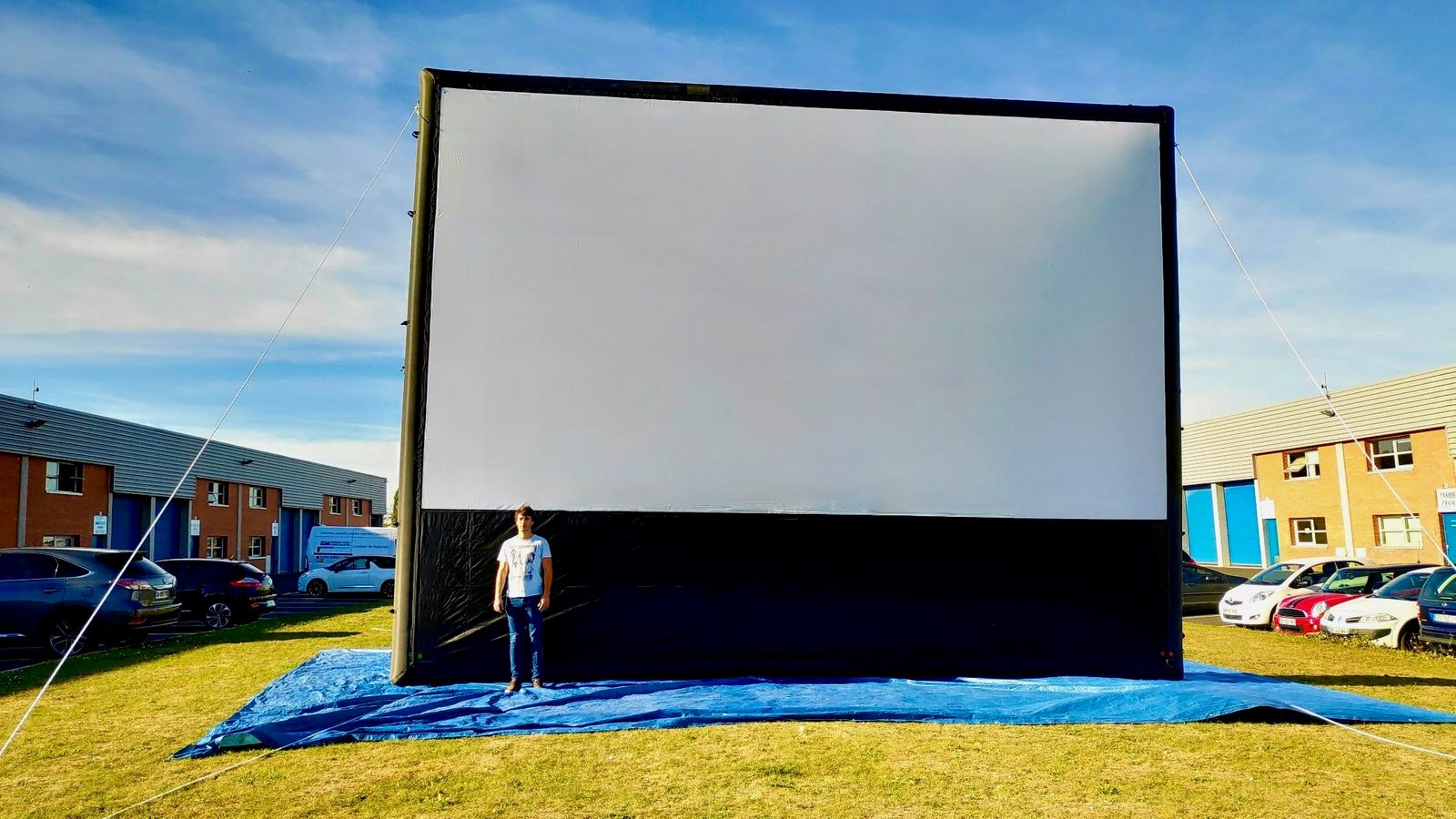 Un homme se tient debout devant un immense écran gonflable blanc et noir installé sur une bâche bleue, entouré de bâtiments industriels et de voitures garées.