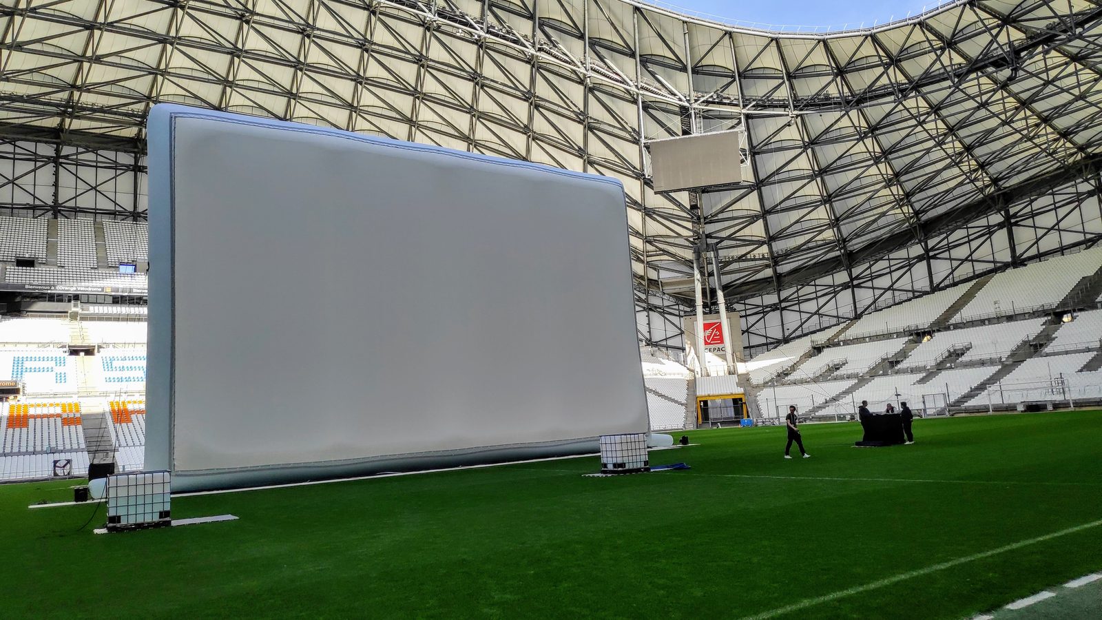 Immense écran gonflable blanc lesté par des cuves d'eau, posé sur la pelouse d'un stade vide sous une charpente métallique.